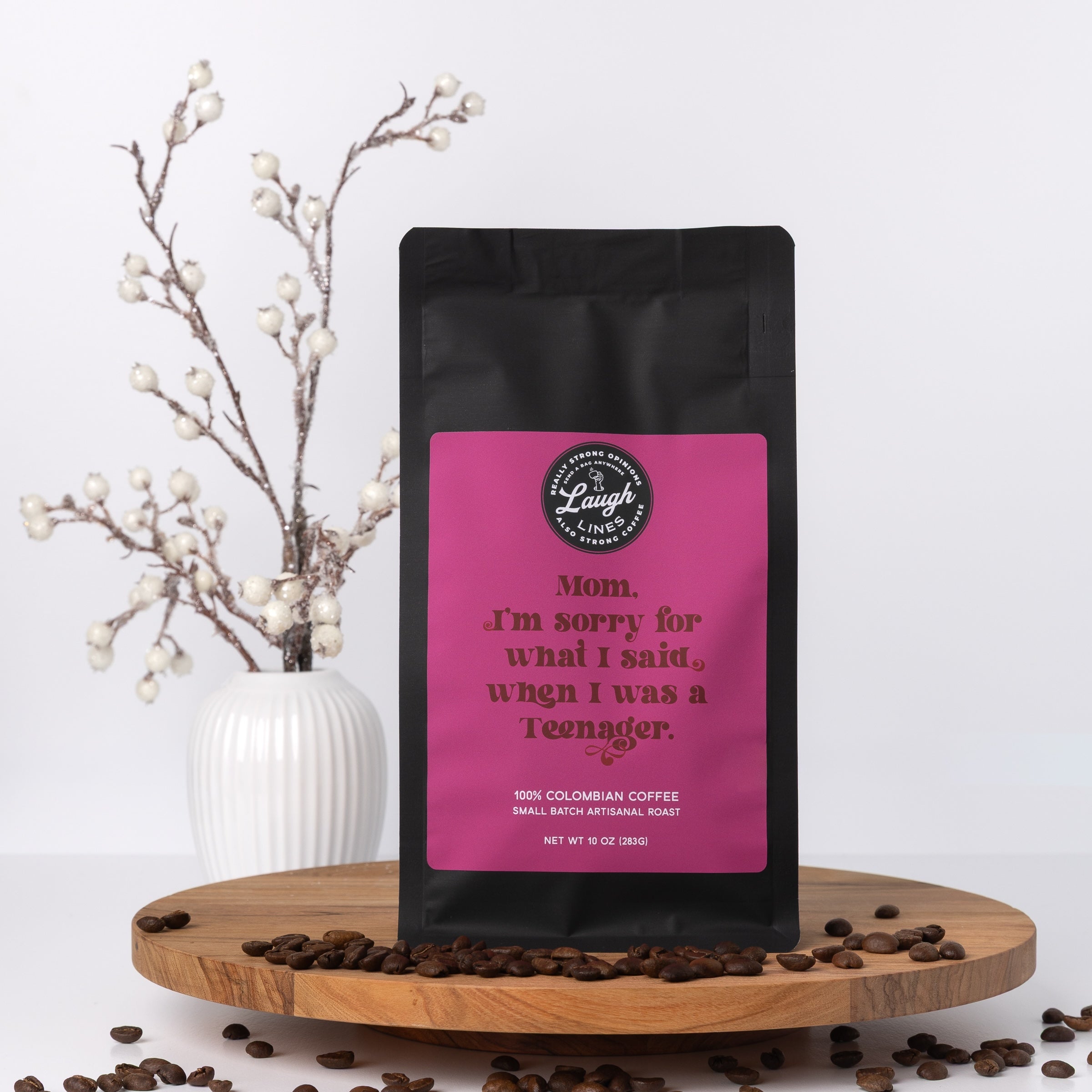 Coffee bag with a pink label  that reads 'Mom. I'm Sorry for What I Said When I Was a Teenage' on a wooden tray with coffee beans and a vase in the background