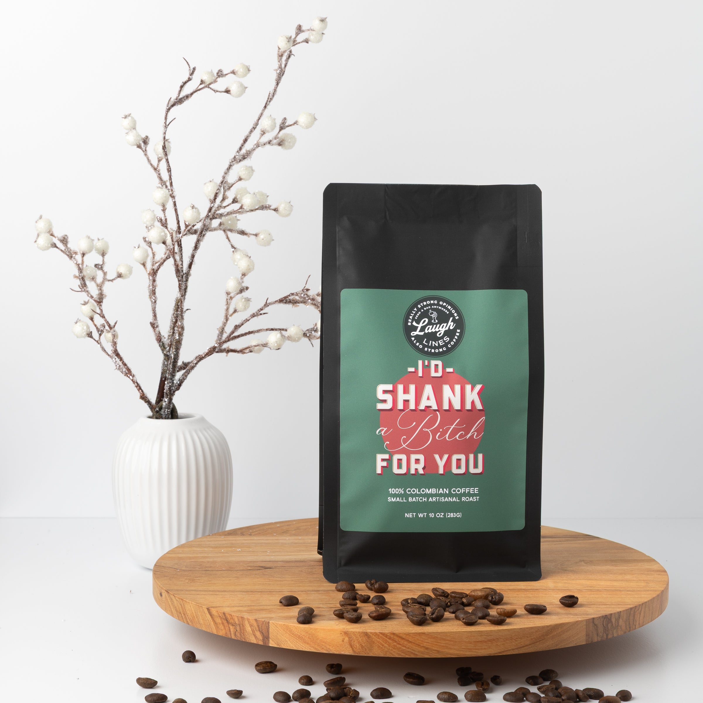 Coffee bag with humorous text that reads' I'd Shank a B*tch for You' on a wooden tray with coffee beans and a vase of branches.