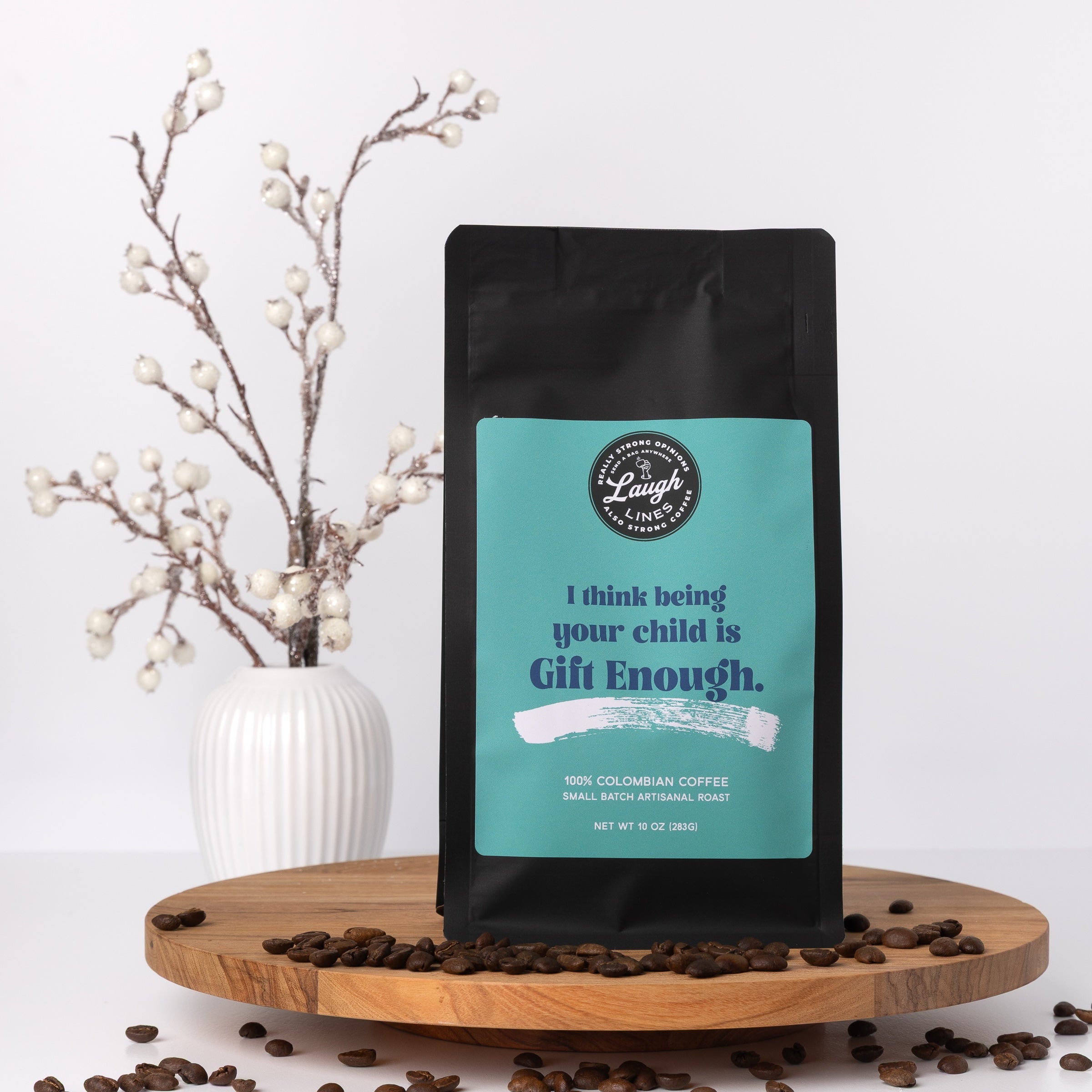 Coffee bag with text on a teal label that reads I think being your child is gift enough on a wooden surface with coffee beans and a decorative plant.