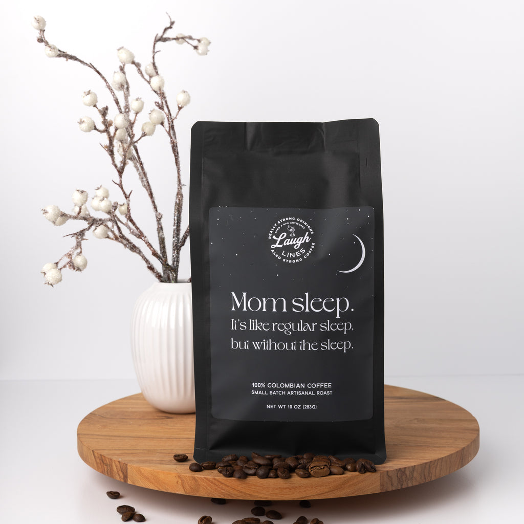 Black coffee bag labeled 'Mom sleep. It's like regular sleep but without the sleep' on a wooden tray with coffee beans and a vase in the background.
