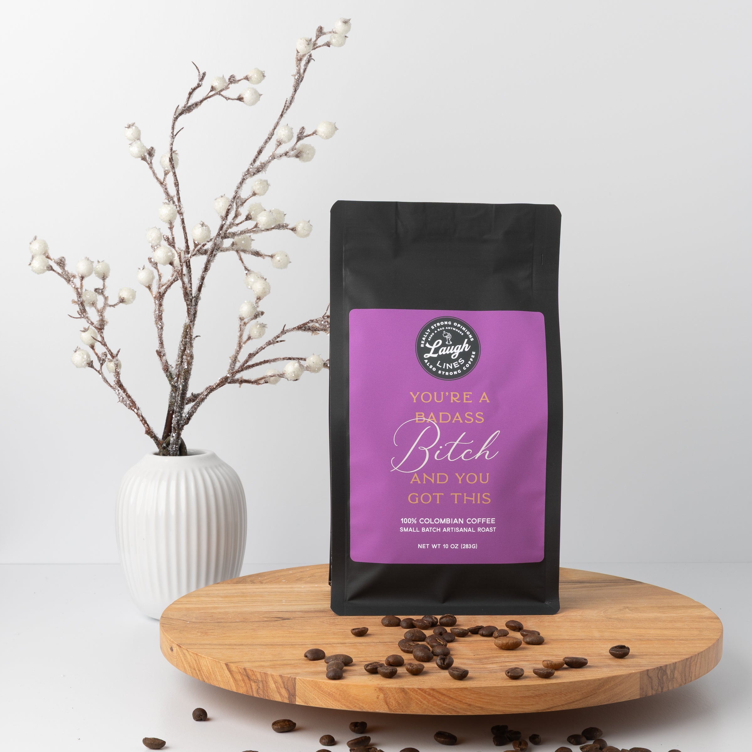 Coffee bag with a purple label that reads, "You're a Badass B*tch and You Got This' on a wooden surface with coffee beans and a vase in the background