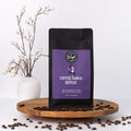 Coffee bag with purple label that says Coffee. Dance. Repeat. on a wooden tray with coffee beans and a vase of branches.