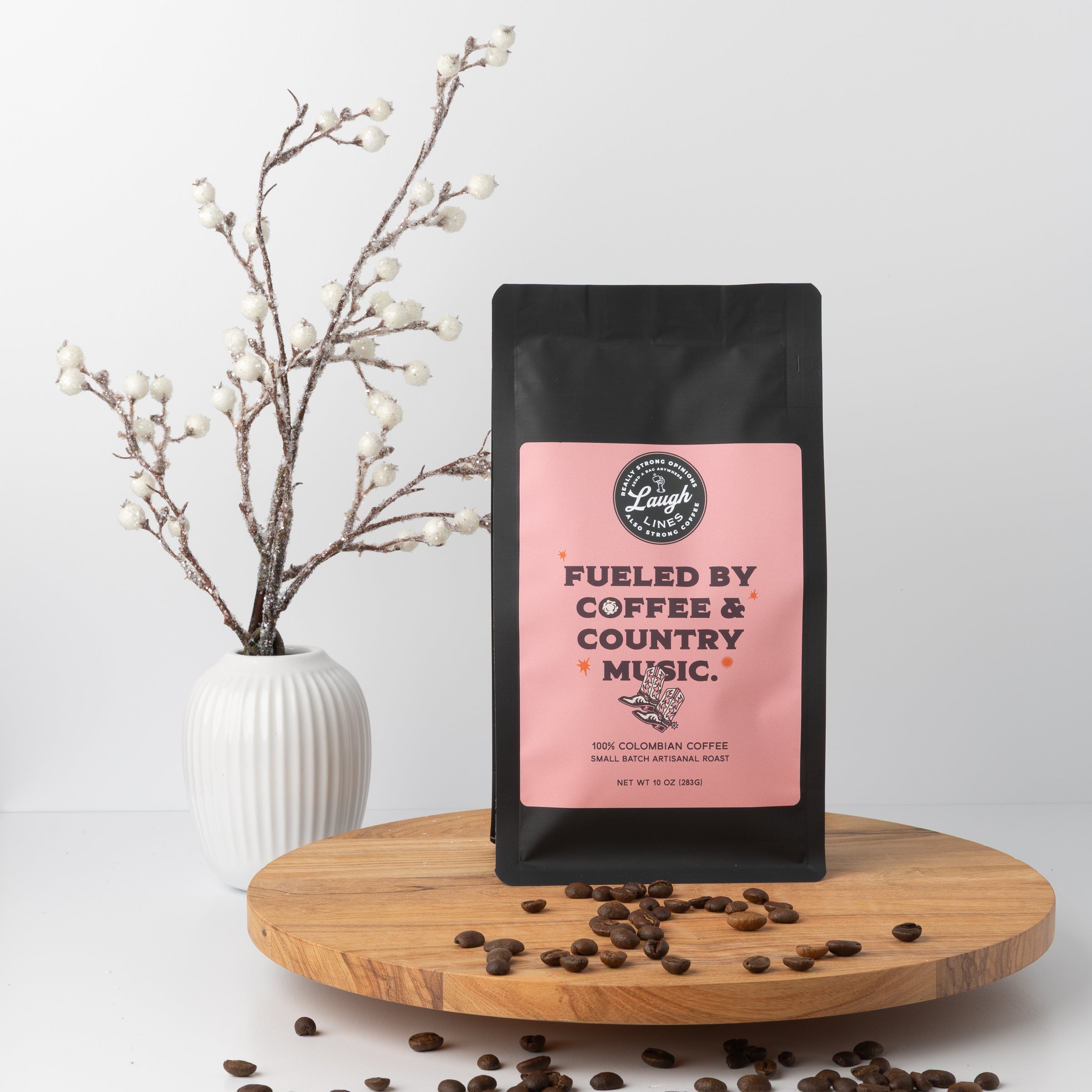Coffee bag with pink label 'Fueled by Coffee & Country Music' on a wooden surface with coffee beans and a vase in the background