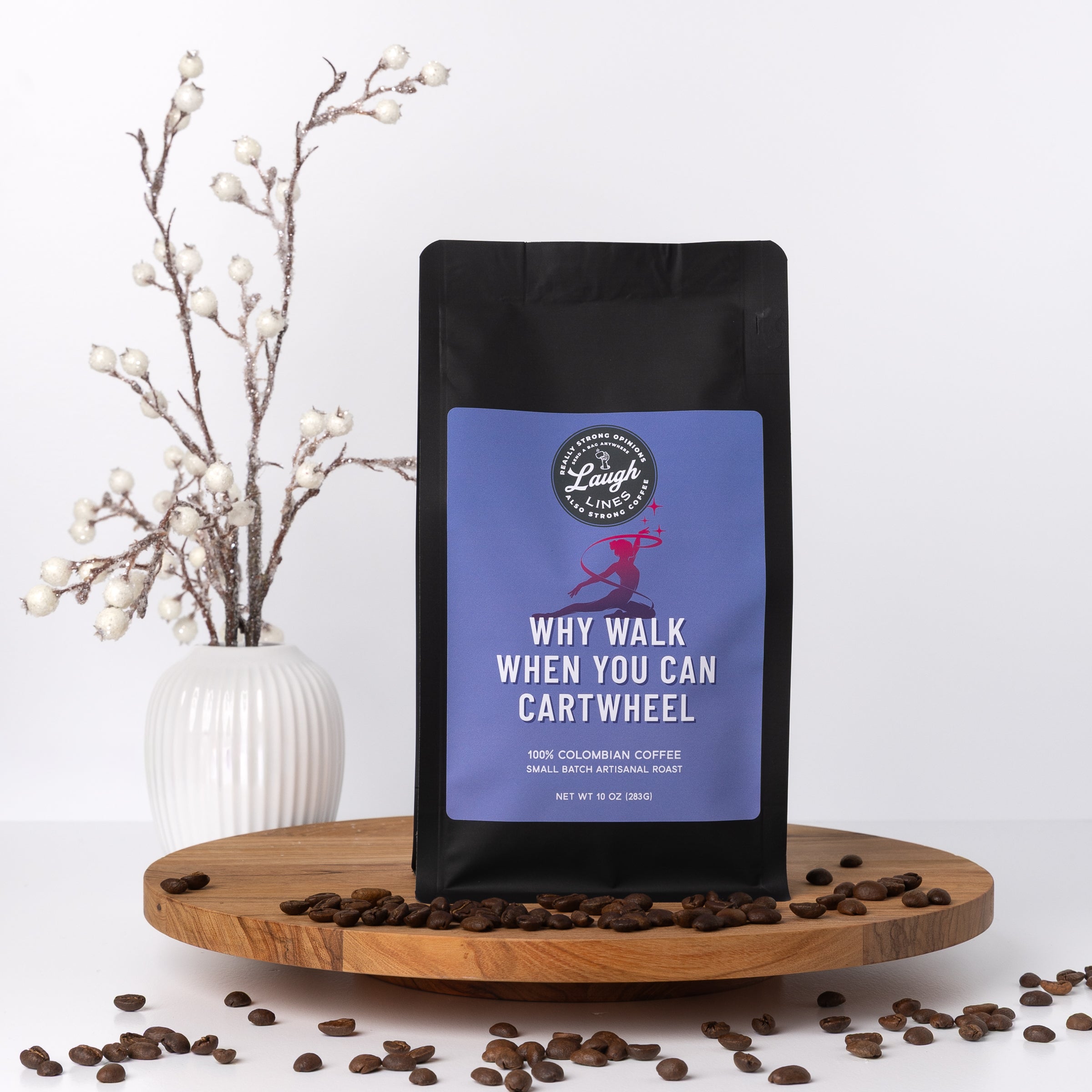 Coffee bag with a humorous label that says. "Wjy Walk When You Can Cartwheel" on a wooden tray with scattered coffee beans, white vase with branches in the background.