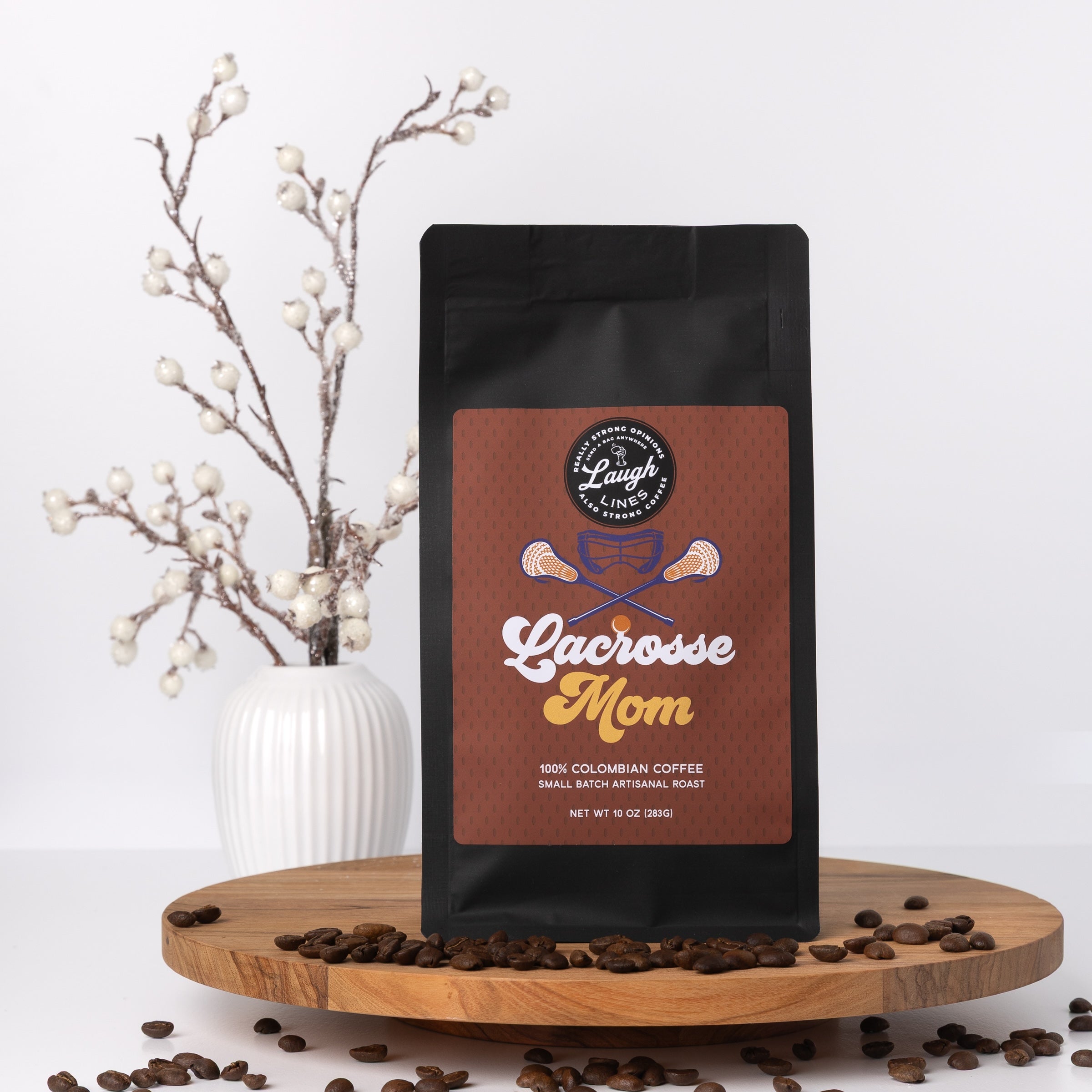Coffee bag labeled 'Lacrosse Mom' on a wooden surface with coffee beans and a decorative vase in the background.