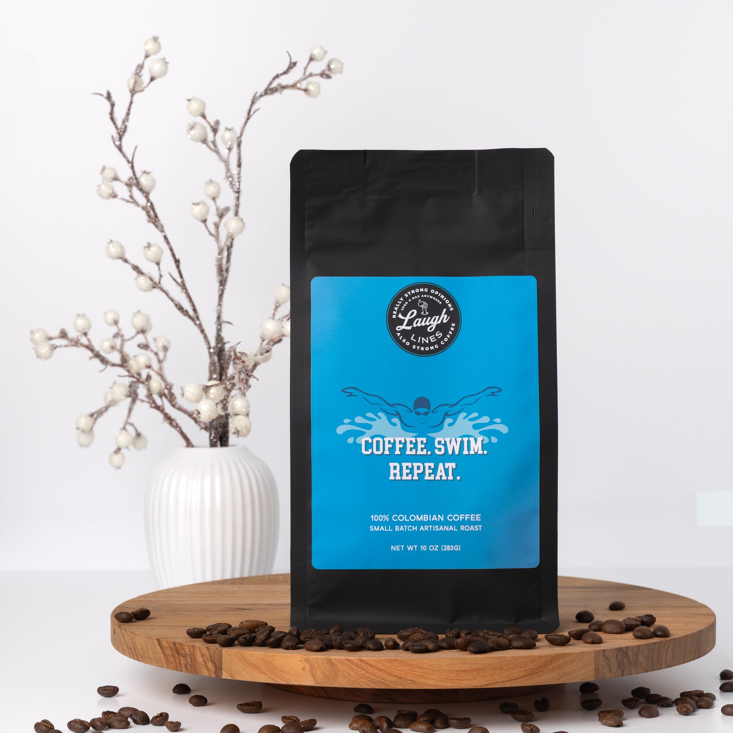 Coffee bag with blue label that says, "Coffee. Swim. Repeat," on a wooden stand with coffee beans and decorative branches.